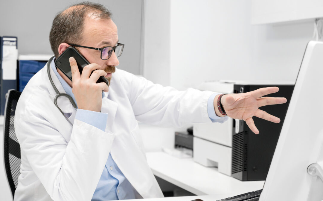 Male doctor gesturing towards a computer monitor while on the phone to illustrate what is practice management